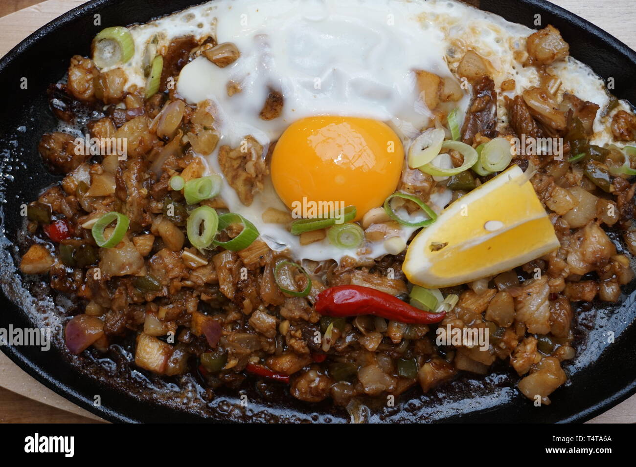 Delicious Pork sisig sizzling mince pork Filipino food with raw egg cooking hot on clay pot tray with lemon slice and fresh red chilies Asian cuisine Stock Photo