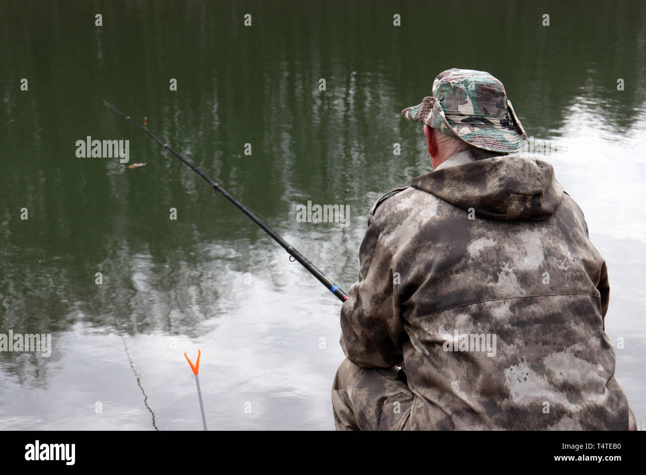 Fisherman in hunting clothes sitting with a fishing rod on the shore. Man angling on the lake in spring, trees are reflected in the water Stock Photo