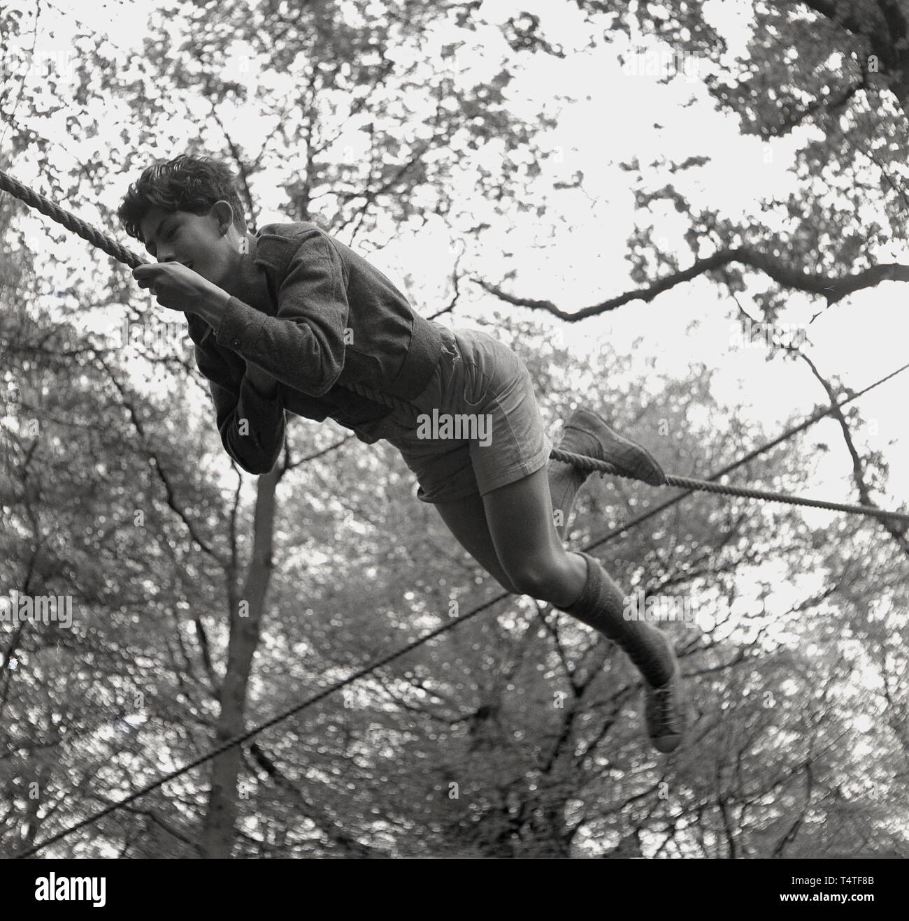 1960s, historical, a young adventure scout lying down on a rope using his hands and feet to manoeuvre himself across a clearing between two trees, England, UK. Young people in the scouts take part in many outdoor adventure activities which help to develop their character and life skills. Stock Photo