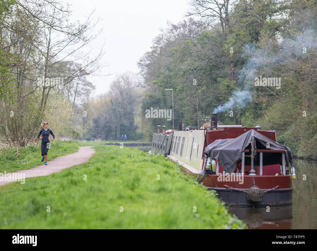 Narrowboat with smoking chimney moored alongside UK canal with female runner (front view) jogging along canal towpath. Stock Photo