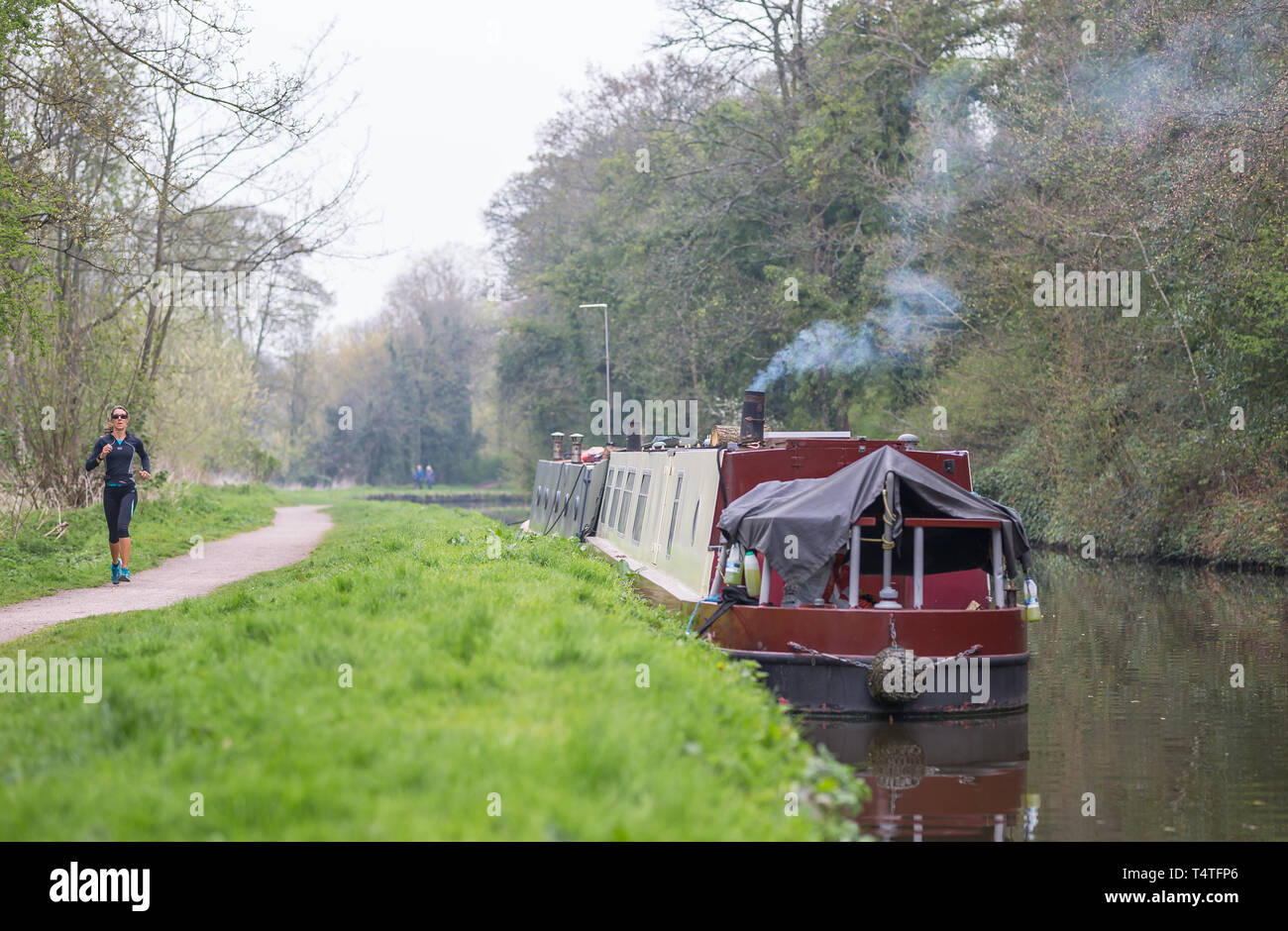 Narrowboat with smoking chimney moored alongside UK canal with female runner (front view) jogging along canal towpath. Stock Photo