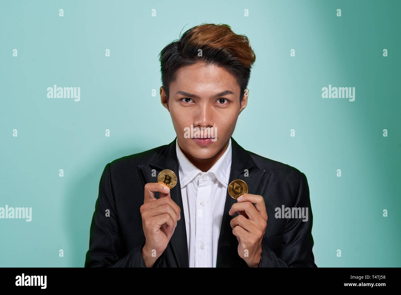 A smiling young man dressed in shirt and jacket showing two golden bitcoin and etherium isolated over gray background Stock Photo