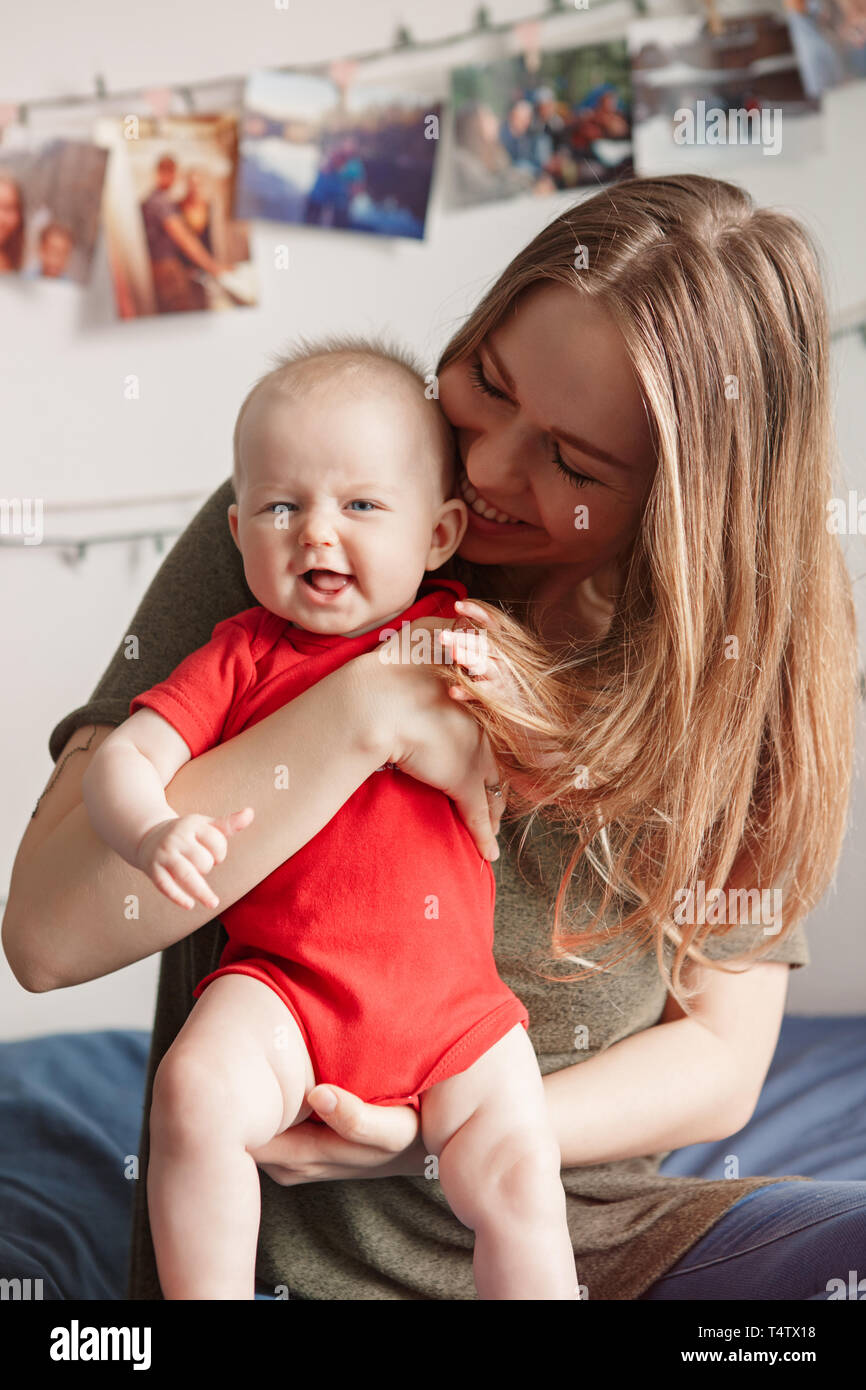 Beautiful white Caucasian woman mother holding cute adorable baby boy girl child sitting on bed in bedroom at home. Authentic candid family lifestyle. Stock Photo