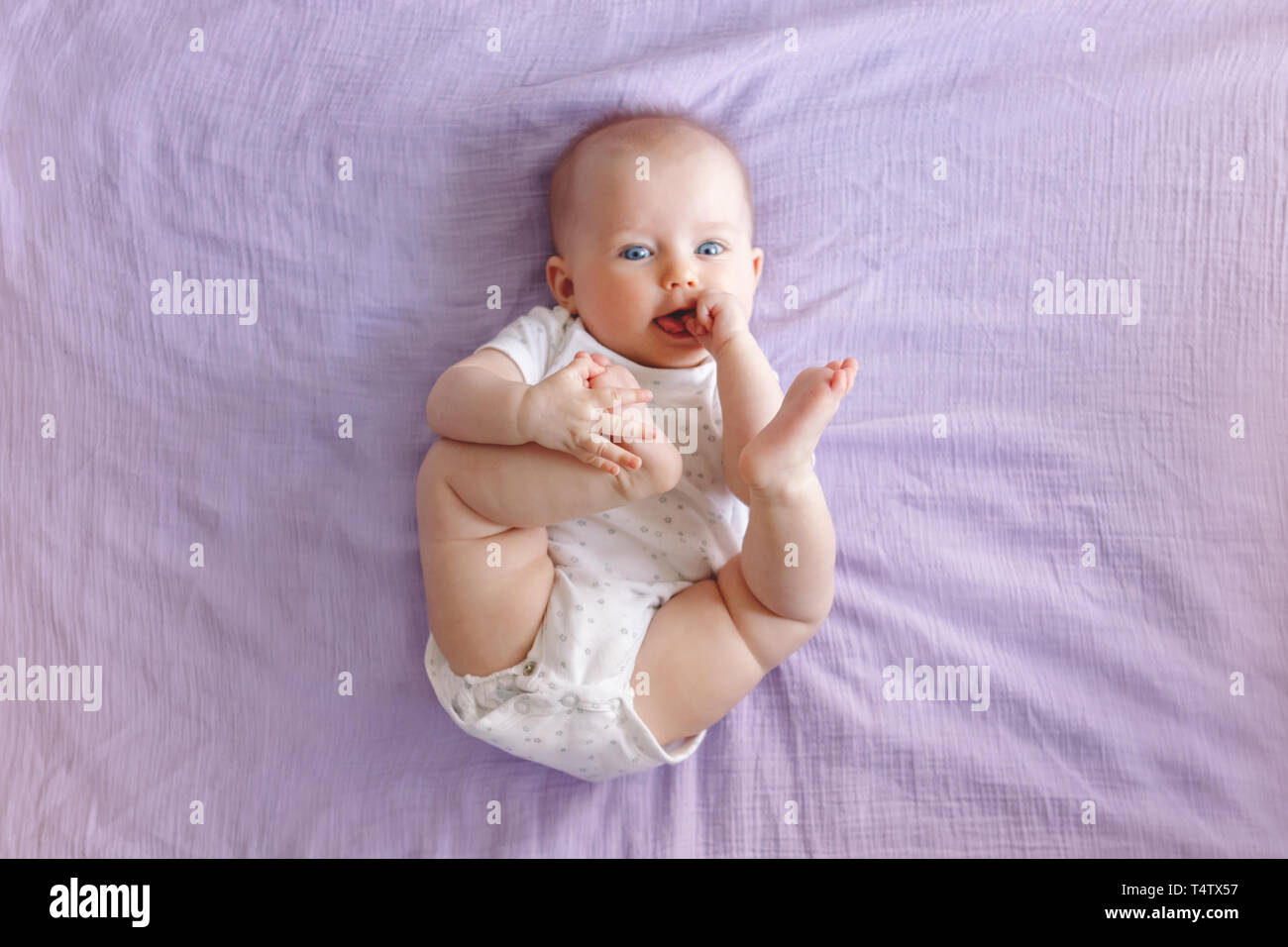 Portrait of cute adorable white Caucasian baby girl boy with blue eyes lying on bed looking at camera and licking sucking fingers fist. View from top  Stock Photo