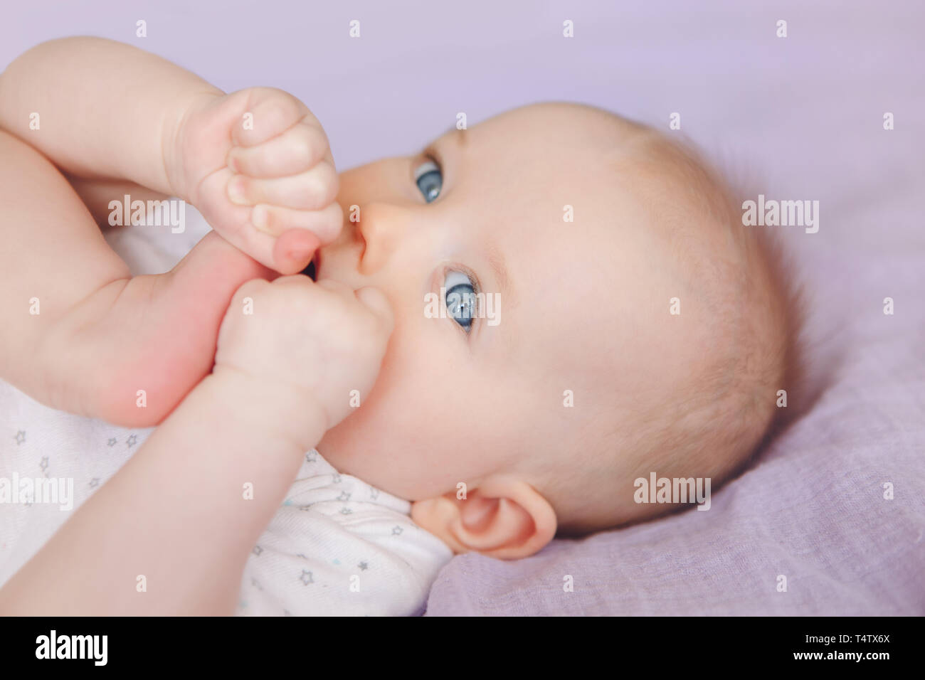 Closeup portrait of cute adorable white Caucasian infant child girl boy with blue eyes lying on bed licking sucking fingers fist. Baby teething. Happy Stock Photo