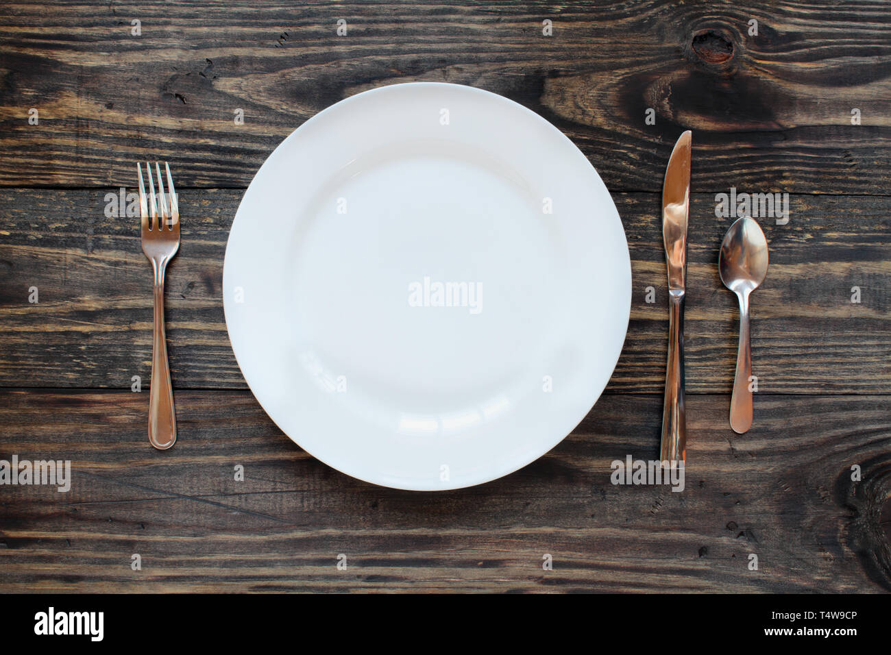 Empty white dinner plate over a rustic wooden table / background with fork, knife and spoon.. Top view. Stock Photo