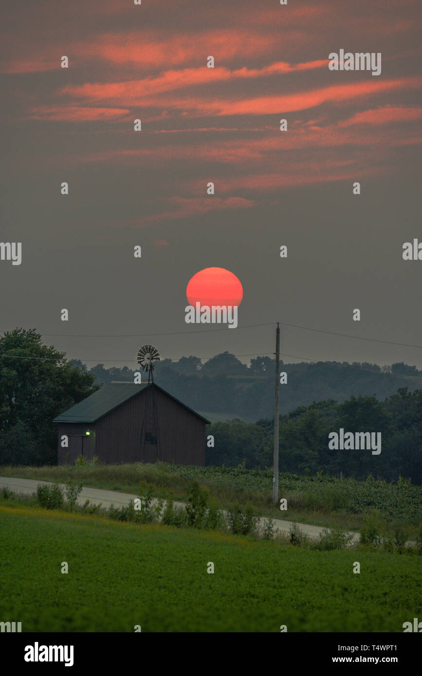 Sultry sunset over rural barn and windmill along countryside road in summer, Monroe, Wisconsin, USA Stock Photo