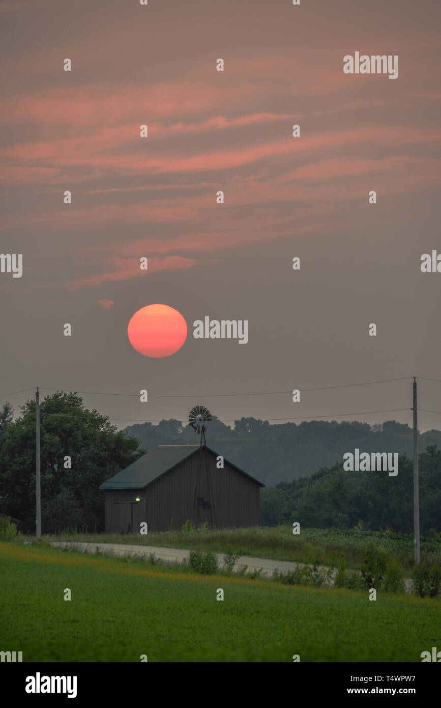 Sultry sunset over rural barn and windmill along countryside road in summer, Monroe, Wisconsin, USA Stock Photo