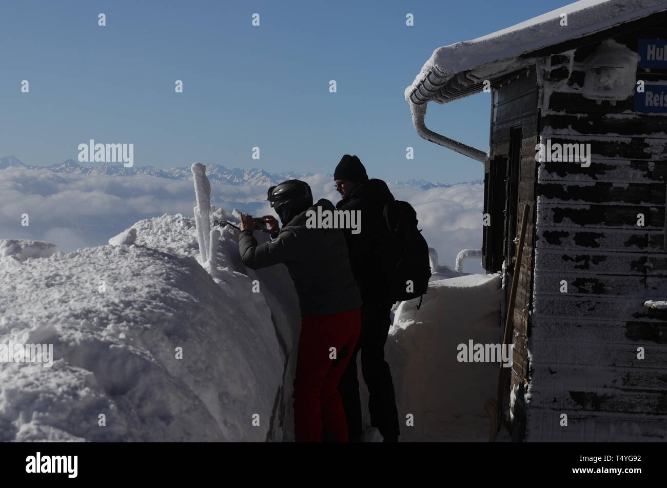 Zugspitze: zwei Besucher stehen hinter einem hohen Schneewall und fotografieren bei klarer Sicht das sonnige und winterliche Alpenanorama. Stock Photo