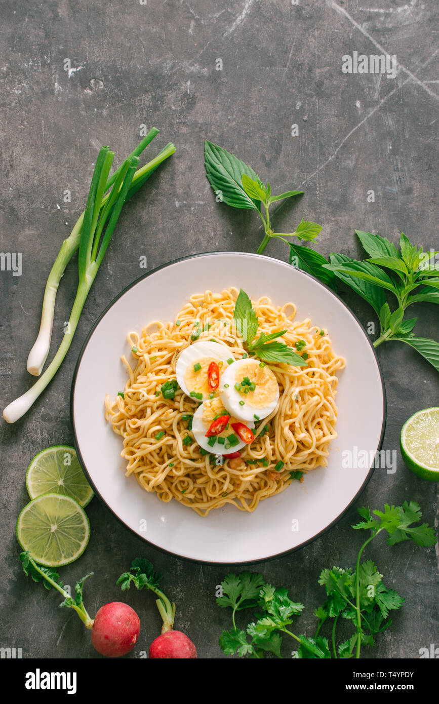 Dry instant noodles put egg with fresh herbs, garnish of cilantro and Asian basil, lemon, lime on dark stone background Stock Photo
