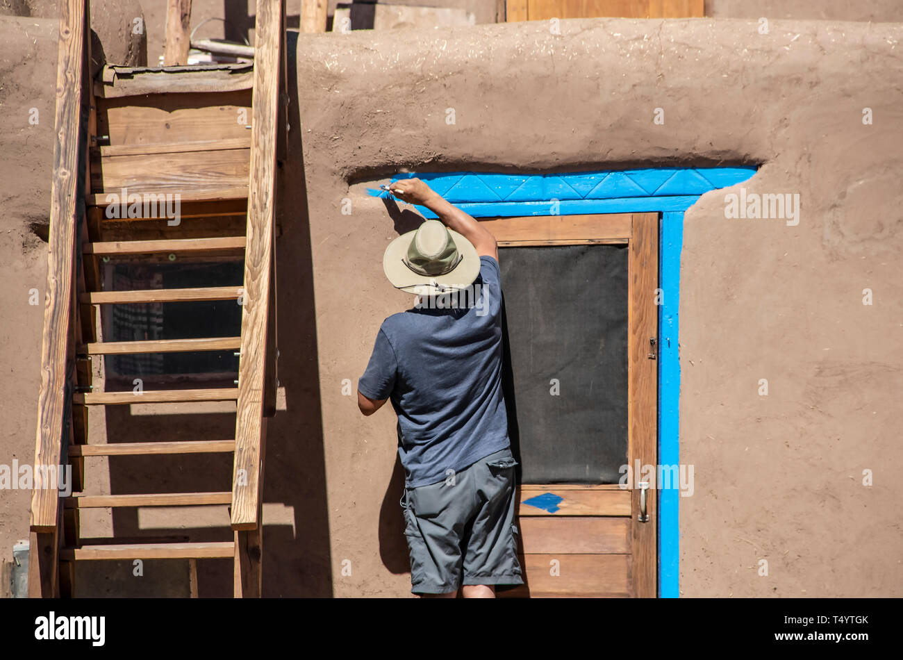 Back of Native American man in shorts and tee-shirt and sun hat painting bright blue-turquoise around the door of a mud adobe pueblo with wooden stair Stock Photo