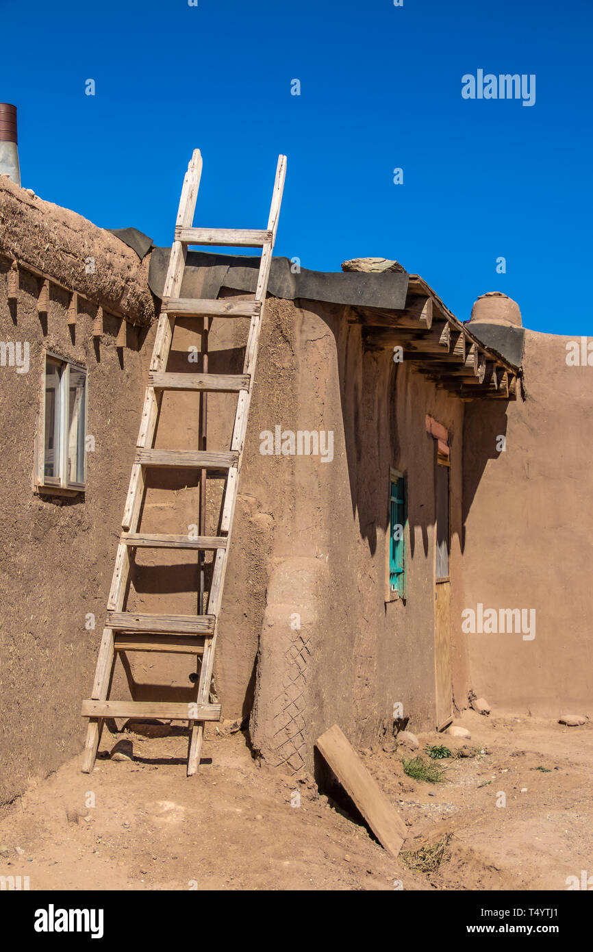 Homemade wooden ladder against side of mud adobe pueblo house where tar paper is being put on roof - with dramatic shadows under intense blue sky Stock Photo