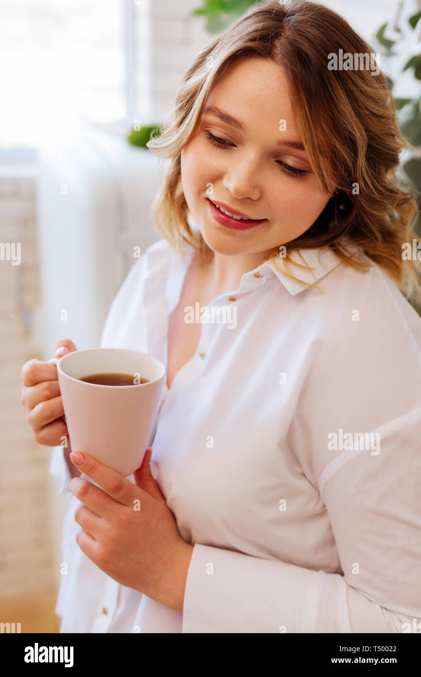 Hot drink. Nice attractive woman holding a cup with hot tea while drinking it in the kitchen