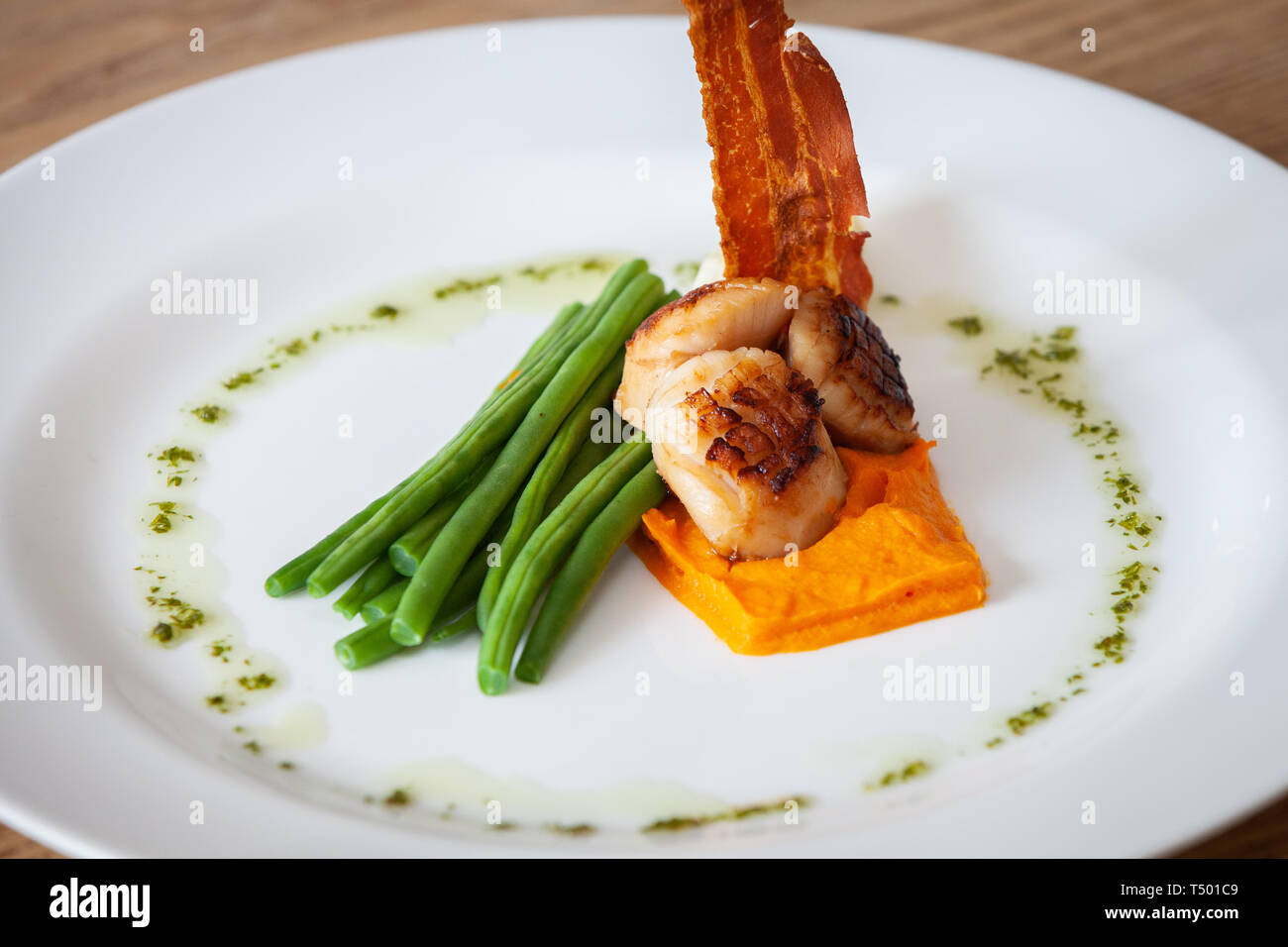 Freshly cooked scallops on a bed of sweet potatoes served with green beans, crispy bacon and salsa verde on a white plate in a restaurant Stock Photo