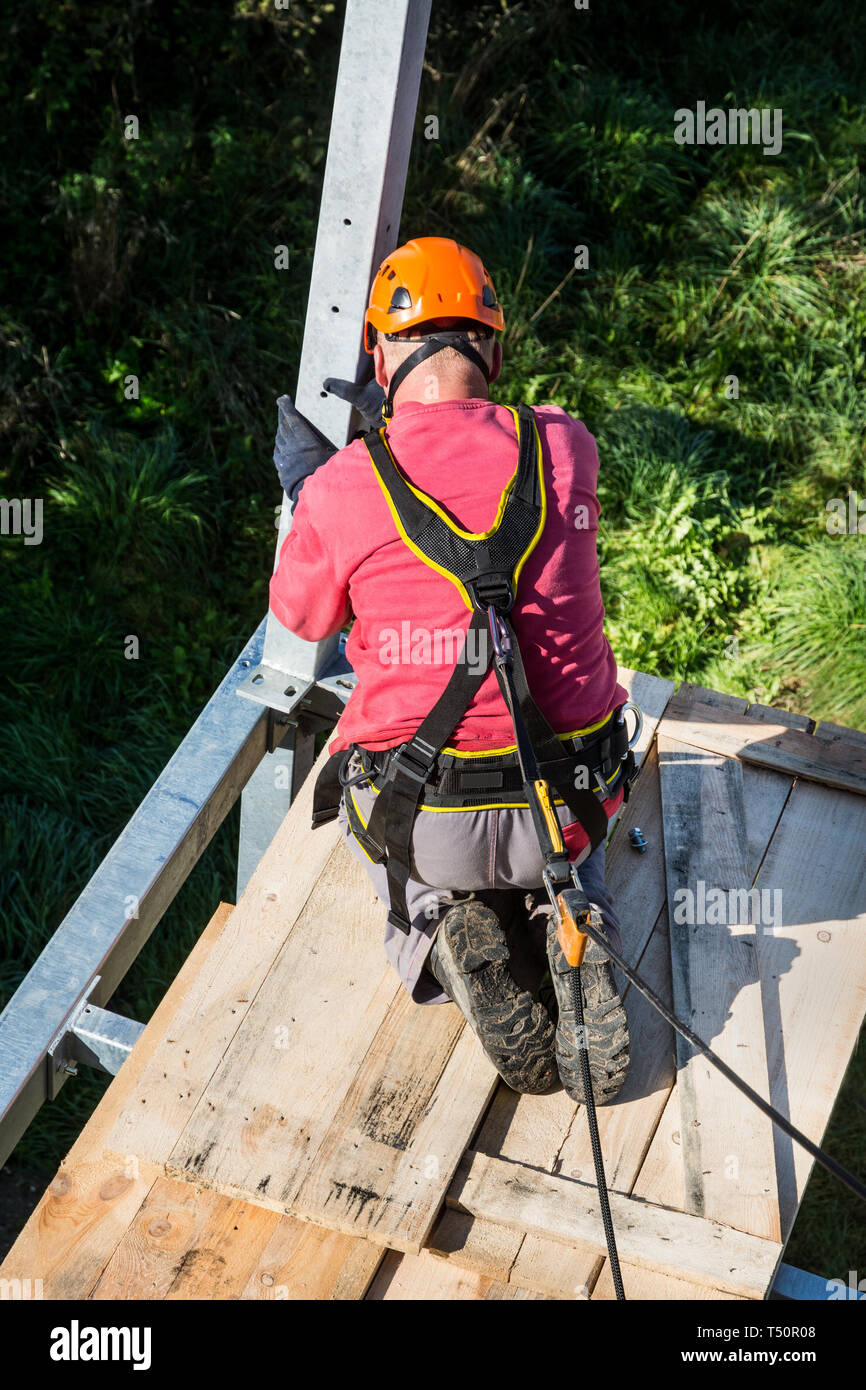 Fall protection. Balcony assembly. Manual worker in seat harness. Workman with orange helmet. Metal framework building. Belaying on rope. Height works. Stock Photo