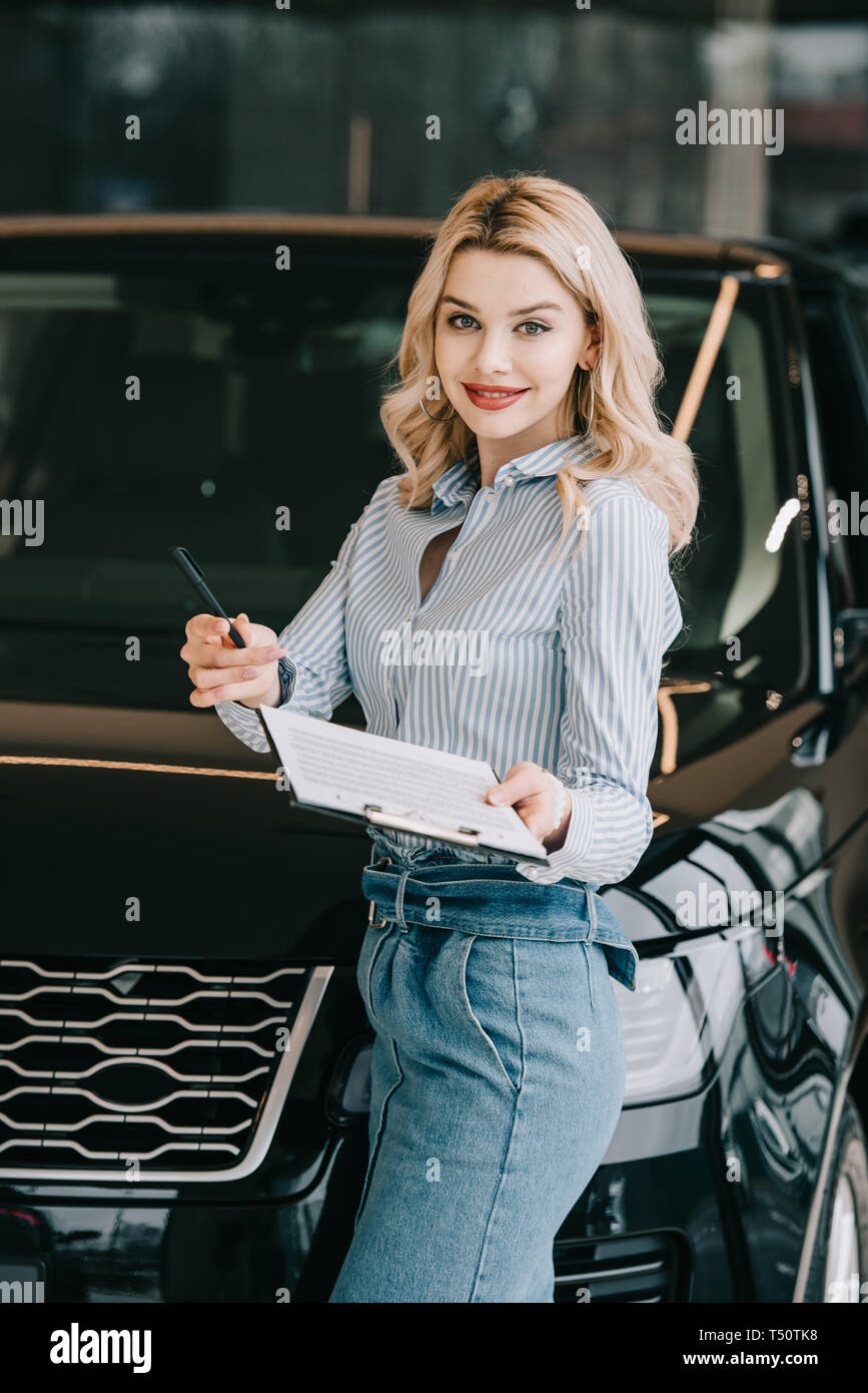 happy blonde car dealer holding clipboard and pen in car showroom Stock ...