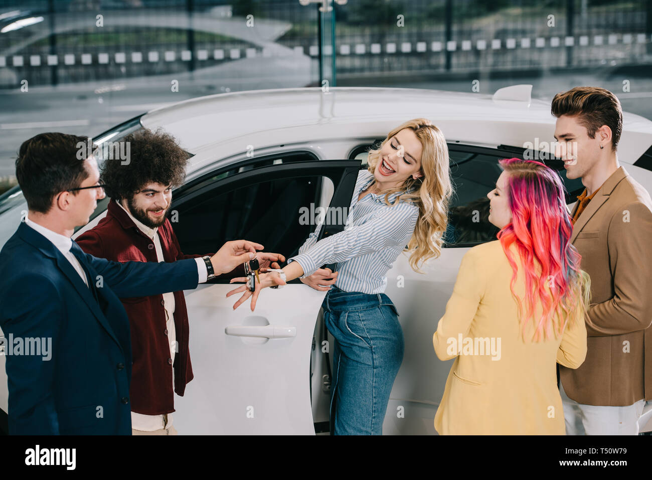 car dealer giving key to happy blonde girl near friends in car showroom