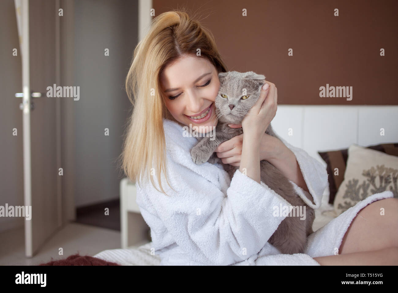 Beautiful young blonde girl in a Terry white robe in her apartment hugs her beloved cat. charming young woman and her pet gray lop-eared cat Stock Photo