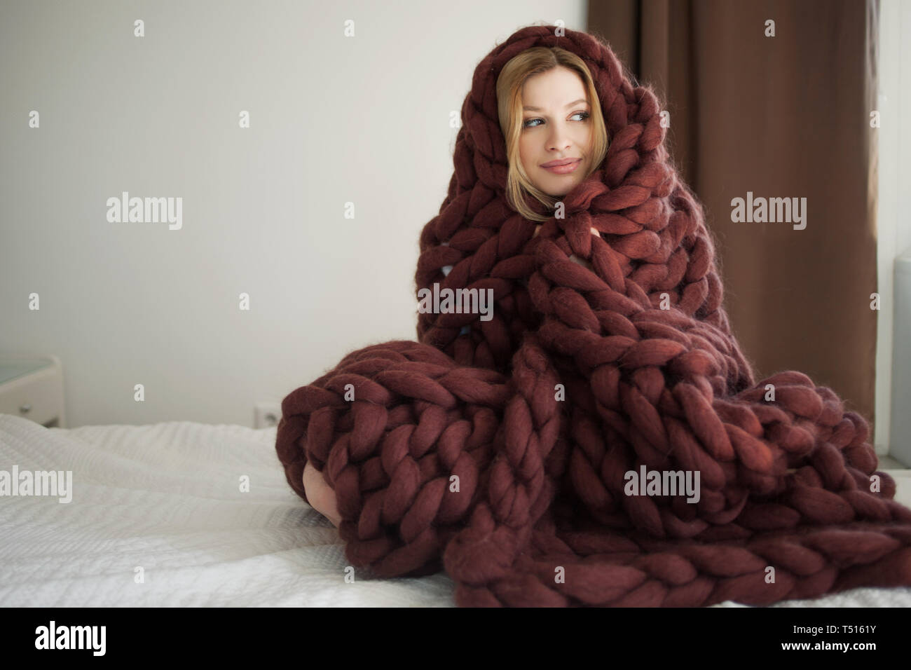 Cute young woman is sitting on the bed wrapped in a big and fluffy ...