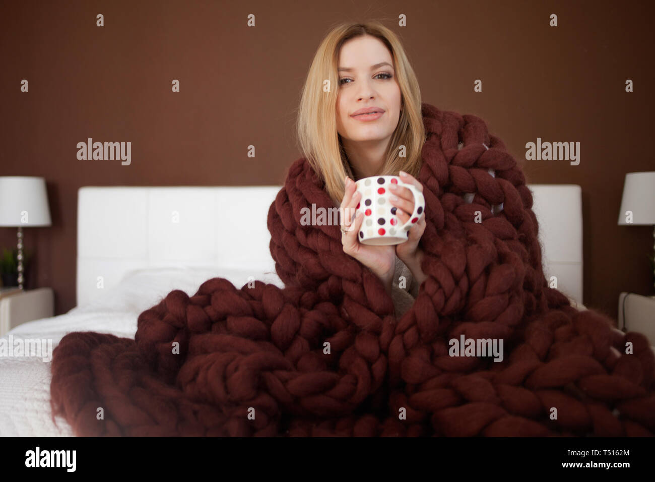 Cute young woman is sitting on the bed wrapped in a big and fluffy ...