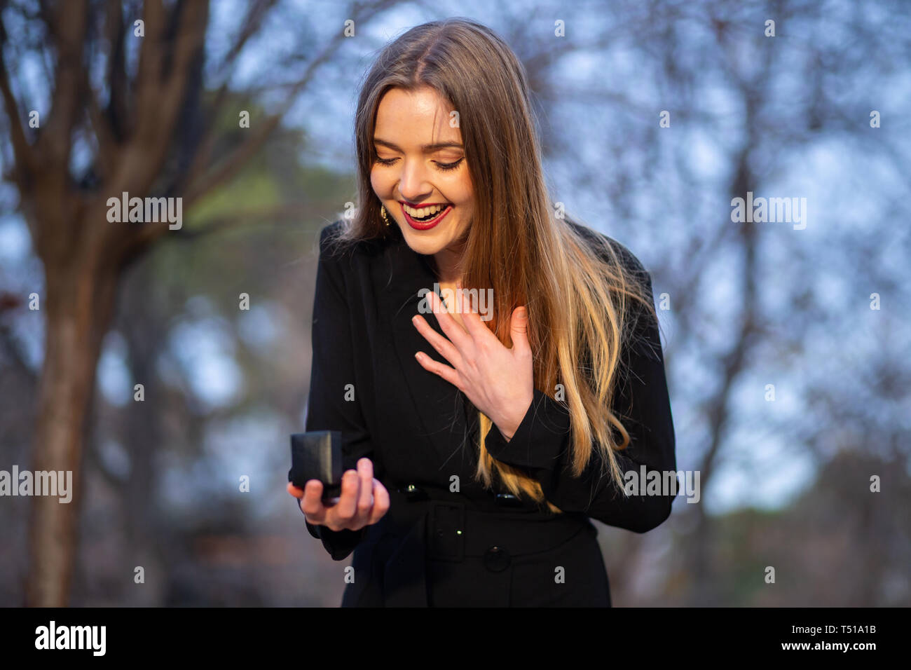 Beautiful young lady in stylish outfit looking at wedding ring in box while standing on blurred background of park Stock Photo