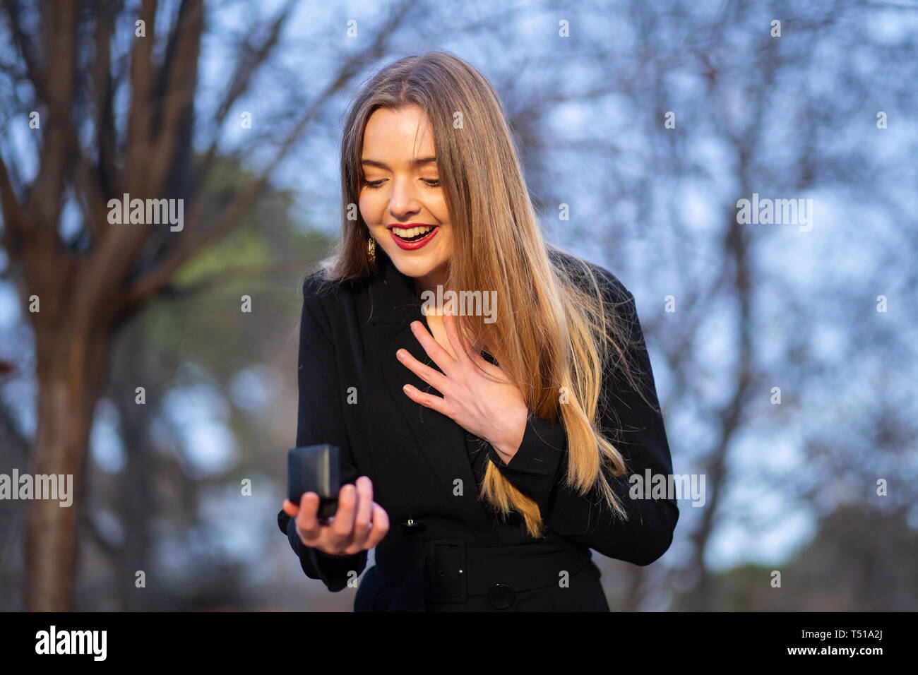 Beautiful young lady in stylish outfit looking at wedding ring in box while standing on blurred background of park Stock Photo