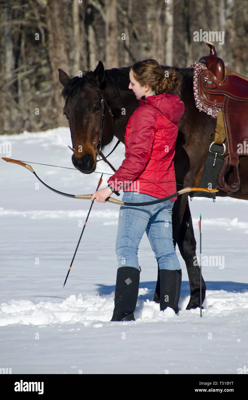 Young woman with saddled quarter horse standing close and holding a bow and arrow, on snowy day in winter, New England Stock Photo