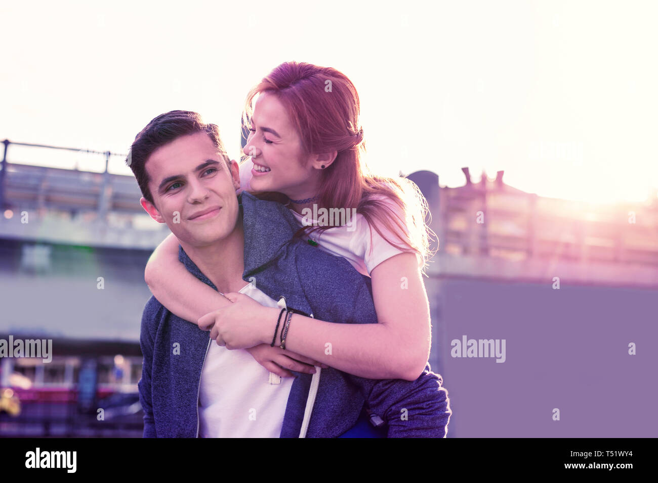 Laughing long-haired ginger girl hanging on her strong handsome boyfriend Stock Photo