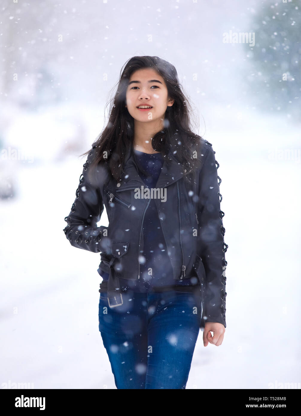 Biracial teen girl wearing black leather jacket and blue jeans walking down middle of street in heavy snowstorm Stock Photo
