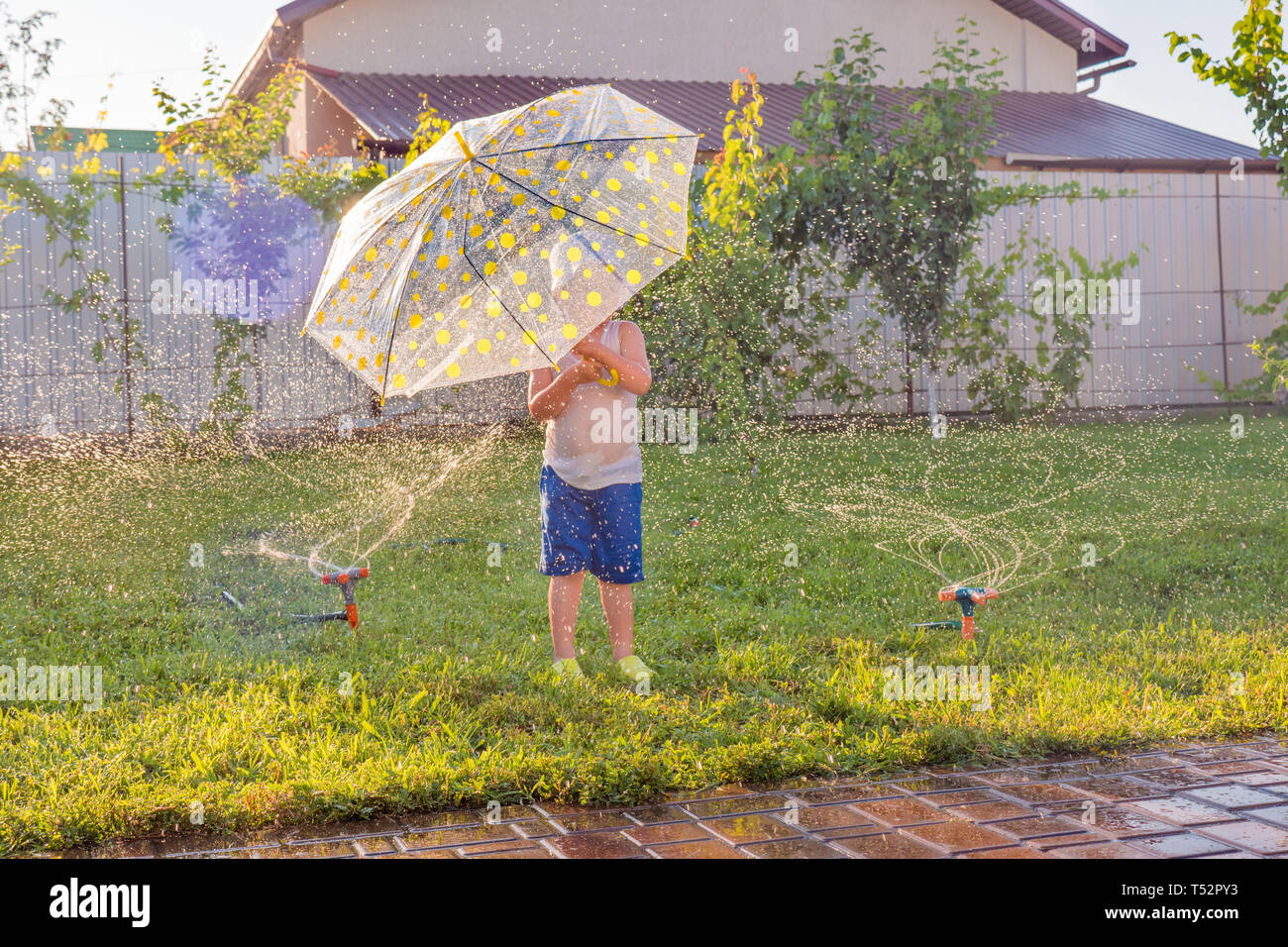 A kid running through water drops of the fontain on front yard with automatic watering system. Exciting games outdoor. Boy with umbrella playing on Stock Photo
