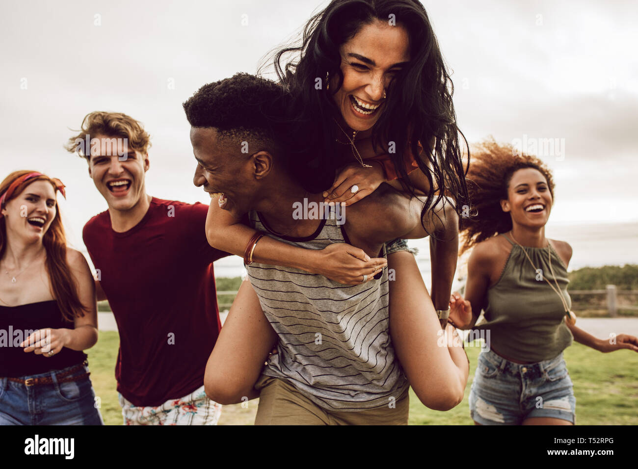 Smiling group of friends enjoying on weekend. Multi-ethnic men and women having fun outdoors. Man carrying woman in his back and running with friends  Stock Photo