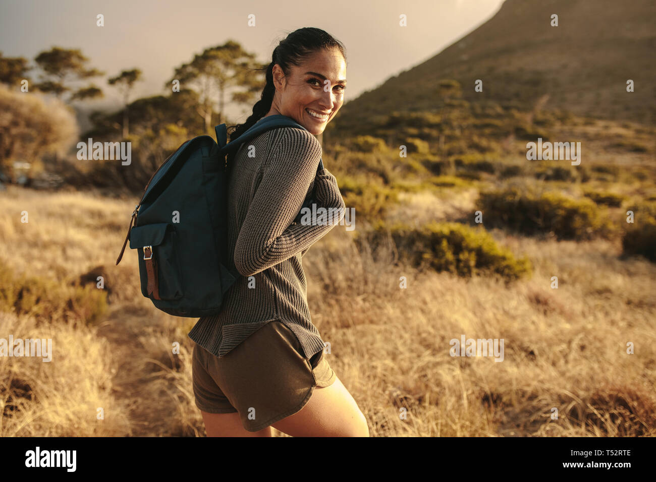 Woman with backpack walking up the country trail and looking over her shoulder and smiling. Stock Photo