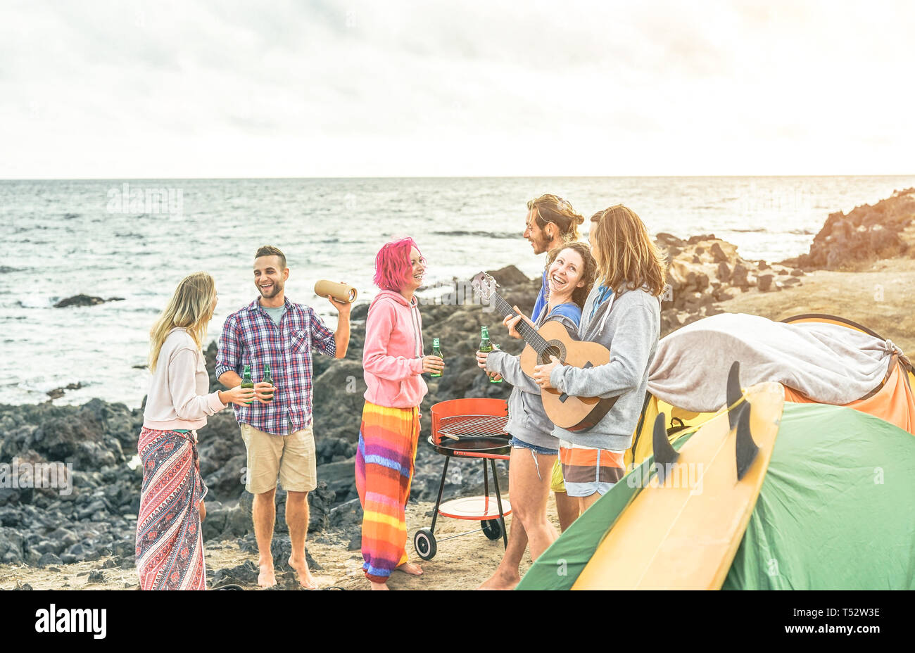 Group of happy friends celebrating drinking beer and playing guitar while camping with tent - Surfers people having fun listening music Stock Photo