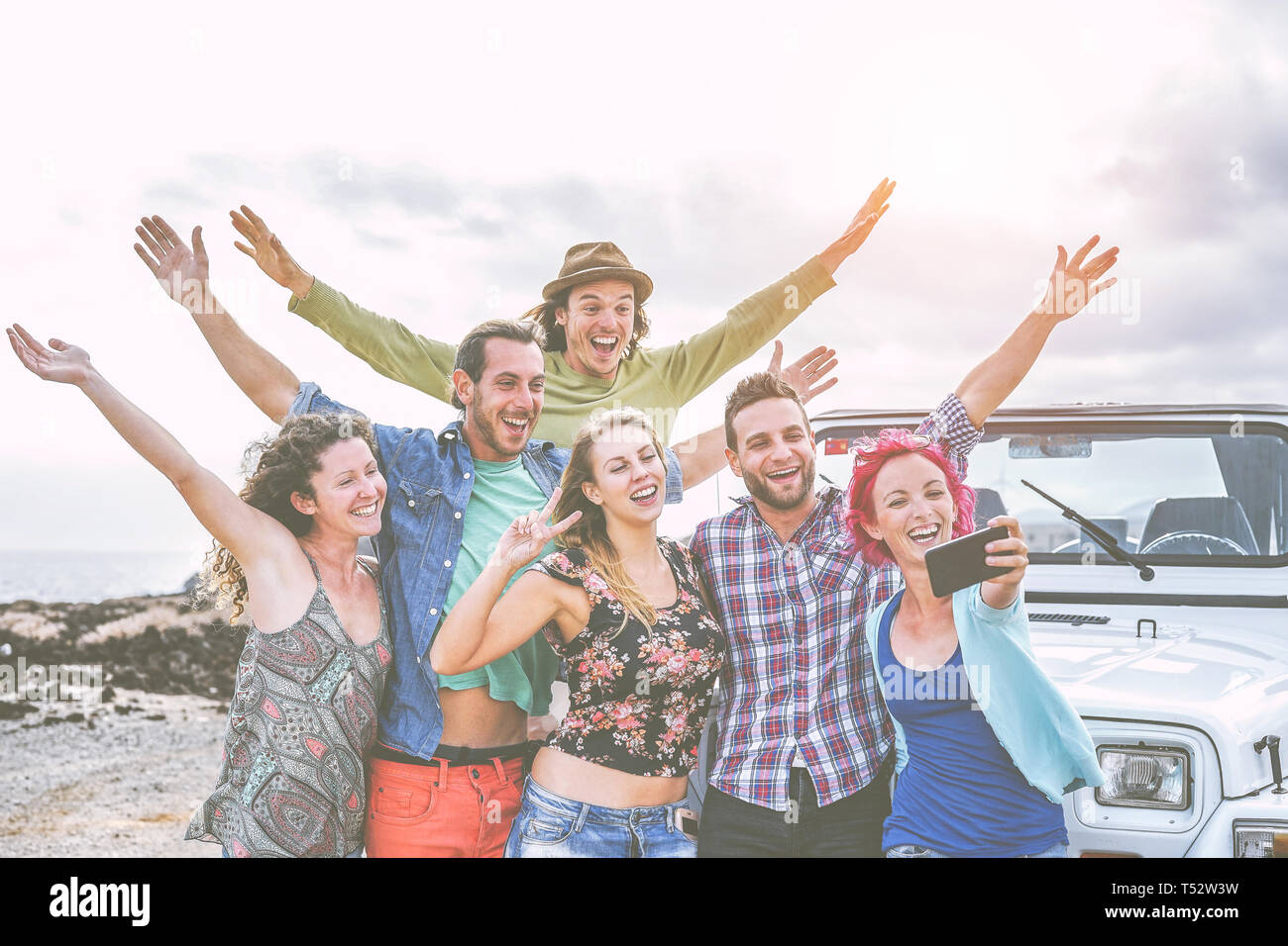 Group of happy friends taking a selfie using mobile smart phone during a road trip with jeep car - Travel people having fun spreading hands up Stock Photo