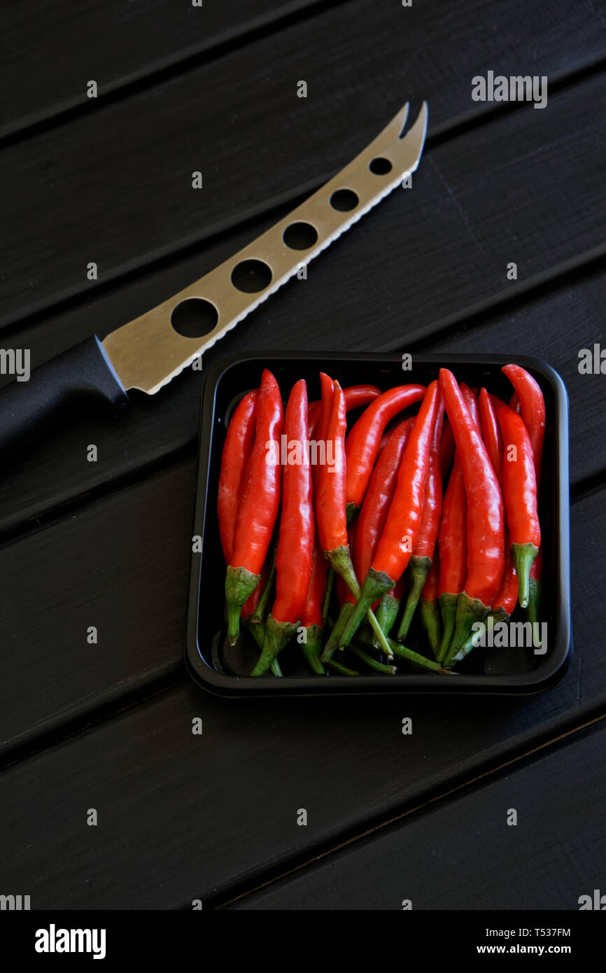 A box of red chilli pods and a knife for cheese and vegetables lie on a black wooden surface. Daylight. Vertical picture Stock Photo