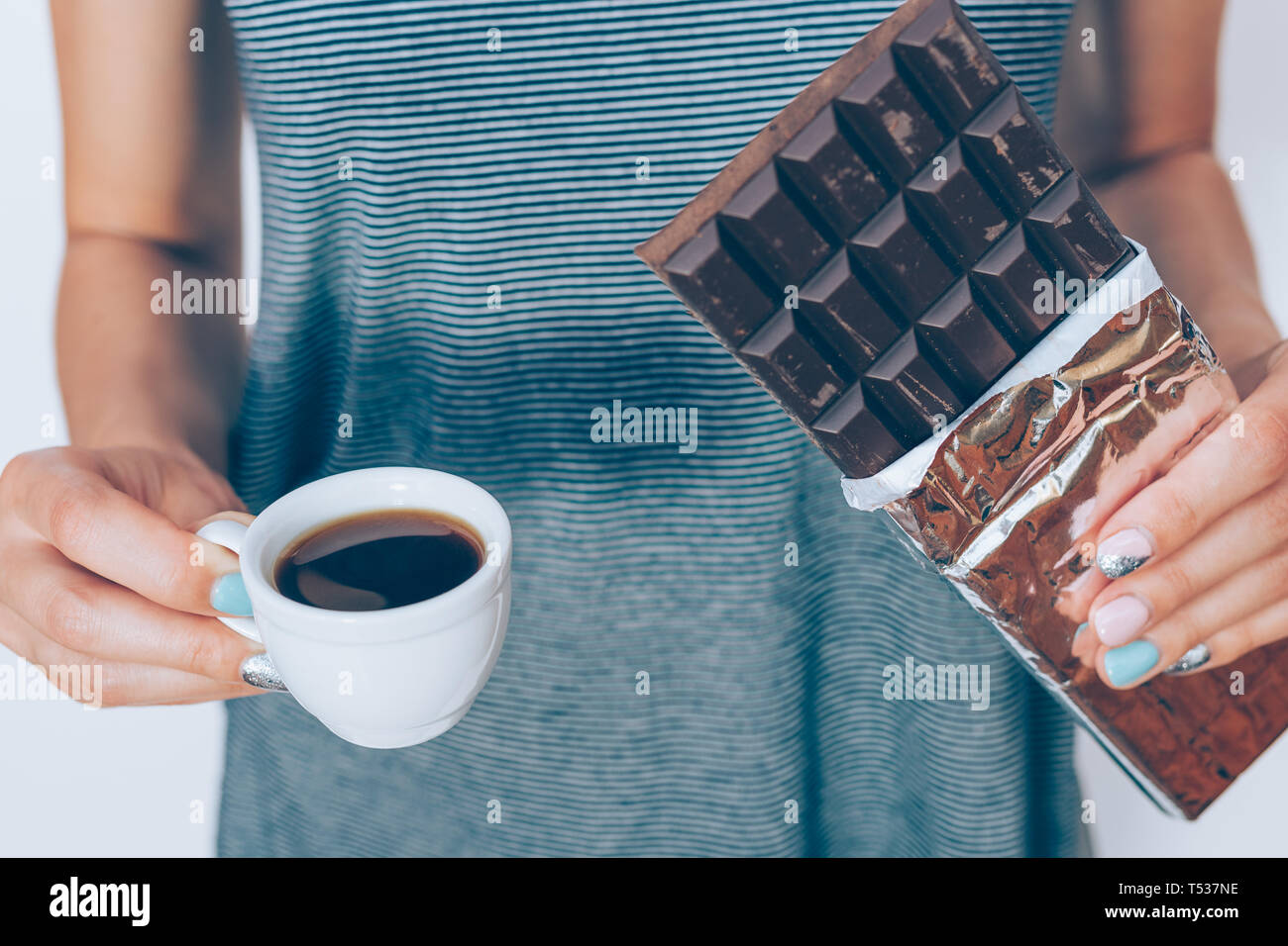 Female's hands holding unwrapped chocolate bar in foil and cup of fresh espresso, close-up. Close-up young woman with cocoa dessert and coffee. Stock Photo