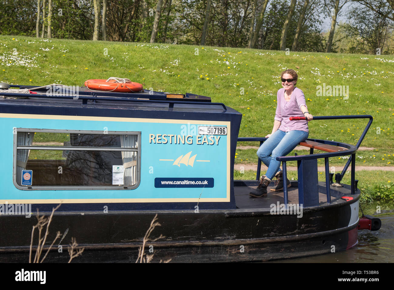 Front view of isolated smiling female lady at rear end of narrowboat, steering the boat, travelling at leisurely pace down UK canal in spring sunshine. Stock Photo