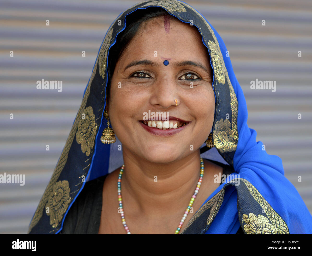 Young Indian Rajasthani woman with blue bindi on her forehead and blue head scarf (dupatta) smiles for the camera. Stock Photo