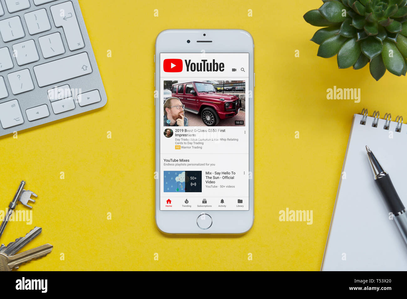 An iPhone showing the YouTube website rests on a yellow background table with a keyboard, keys, notepad and plant (Editorial use only). Stock Photo