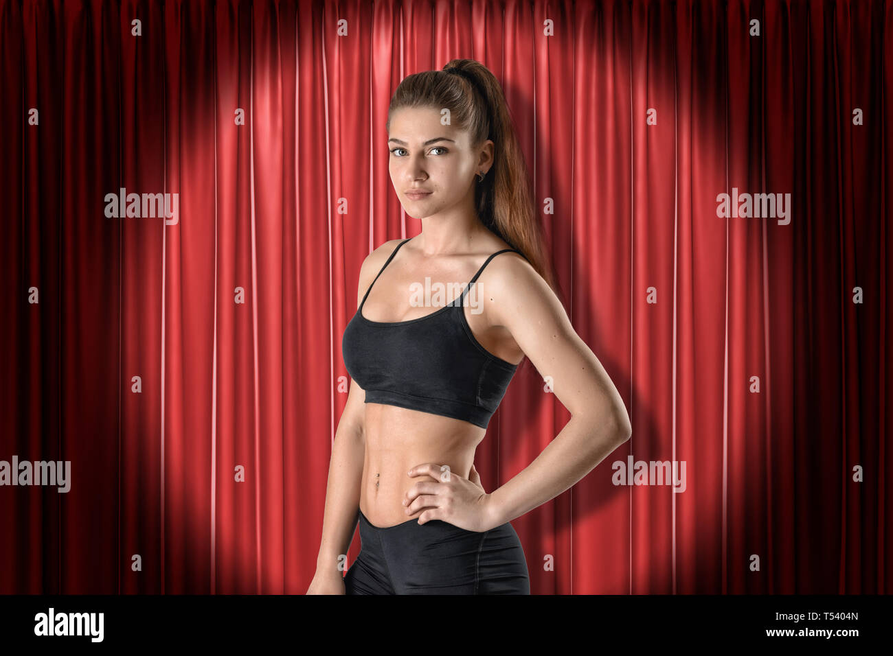 Close-up of young woman in black sport crop top and shorts standing in half-turn with one hand on hip and looking at camera against red curtain. Stock Photo