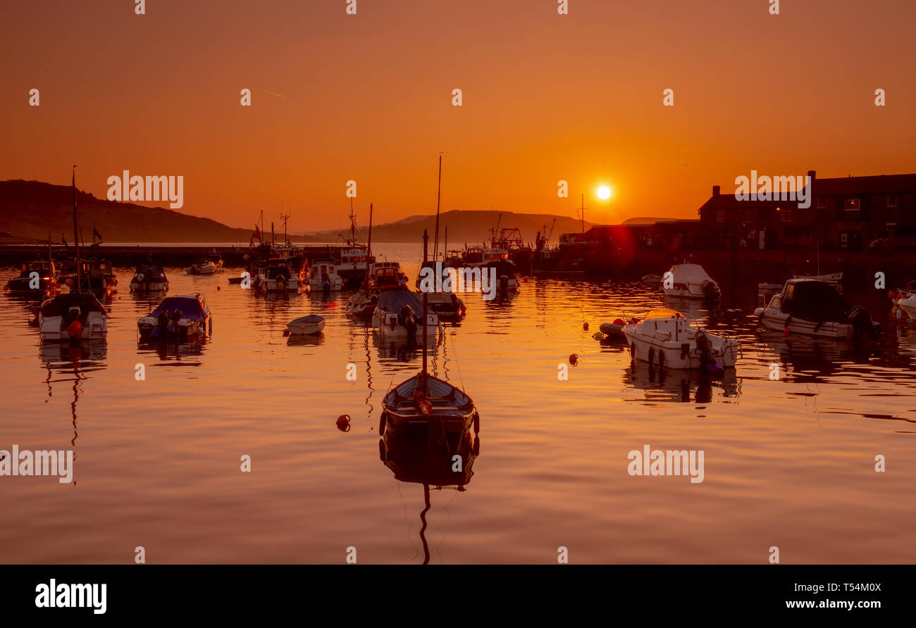 Lyme Regis, Dorset, UK. 21st April 2019. UK Weather: The boats in the historic Cobb Harbour are silhouetted against vibrant orange sky as the sunrises on a gloriously sunny Easter Sunday. Credit: Celia McMahon/Alamy Live News Stock Photo