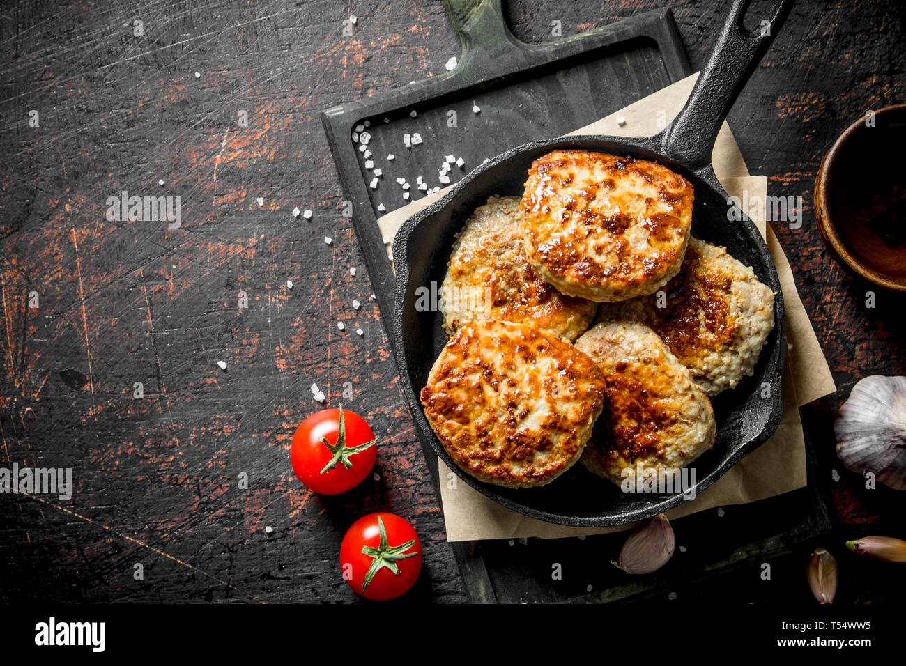 Cutlets in the pan on the paper with garlic and tomatoes. On dark rustic background Stock Photo