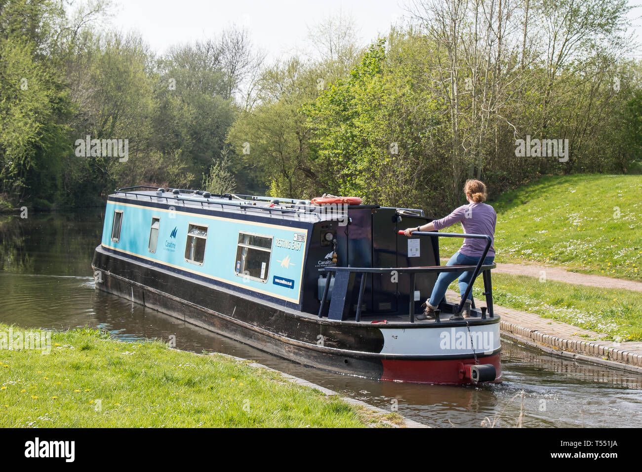 Rear view of single female at rear end of narrowboat, steering the long boat, travelling at a leisurely pace, down UK canal in the spring sunshine. Stock Photo