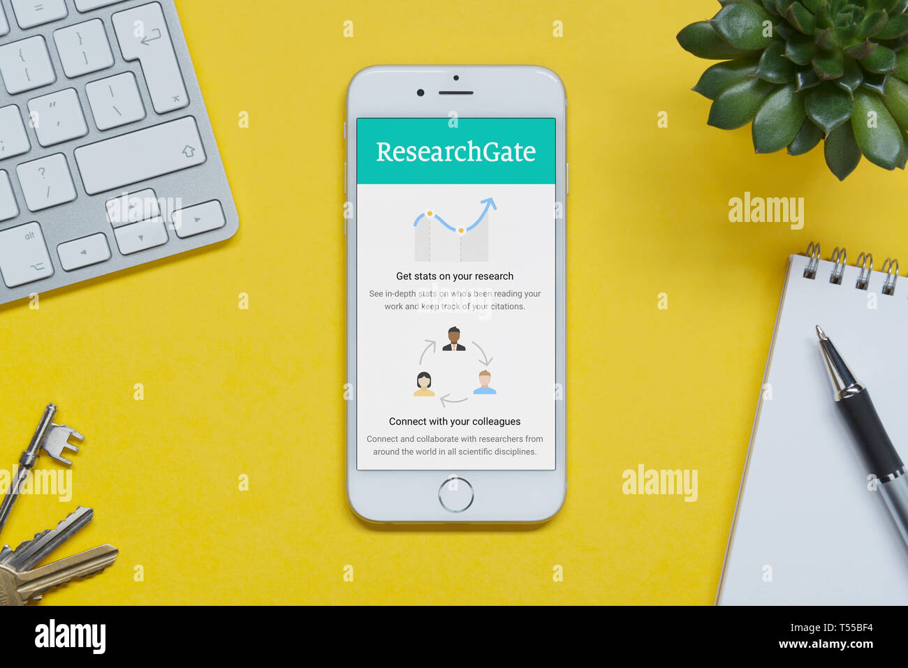 An iPhone showing the Research Gate website rests on a yellow background table with a keyboard, keys, notepad and plant (Editorial use only). Stock Photo
