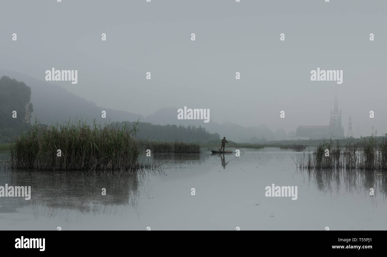 Fisherman on wood boat hunting with long stick on a foggy morning with church in background, Van Long Wetland Nature Reserve, Ninh Binh, Vietnam, Asia Stock Photo
