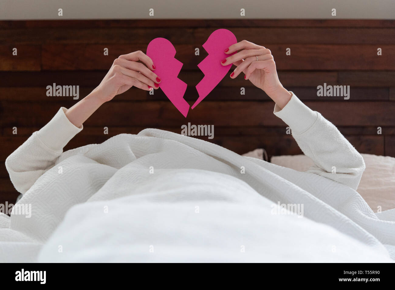 Woman holding aloft pink broken heart while lying in bed Stock Photo