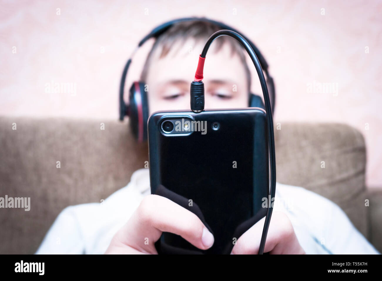 Teenage Boy in Headphones Lying On the Sofa and Looking At the Black Smartphone Screen. Listening To Music, Surfing, Learning Online, Playing or Typin Stock Photo