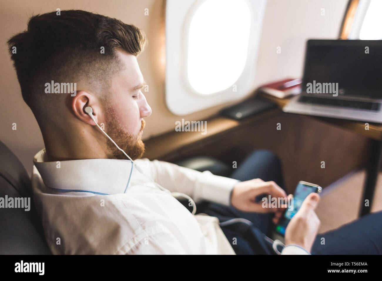 Young attractive and successful businessman is resting and listening to music works while sitting in the chair of his private business plane Stock Photo