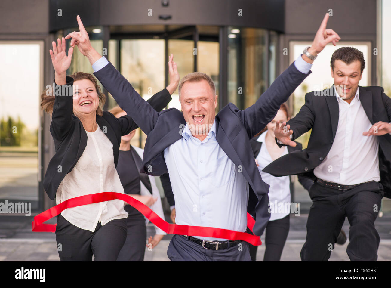Group of happy business people running from office building crossing red ribbon finish line Stock Photo