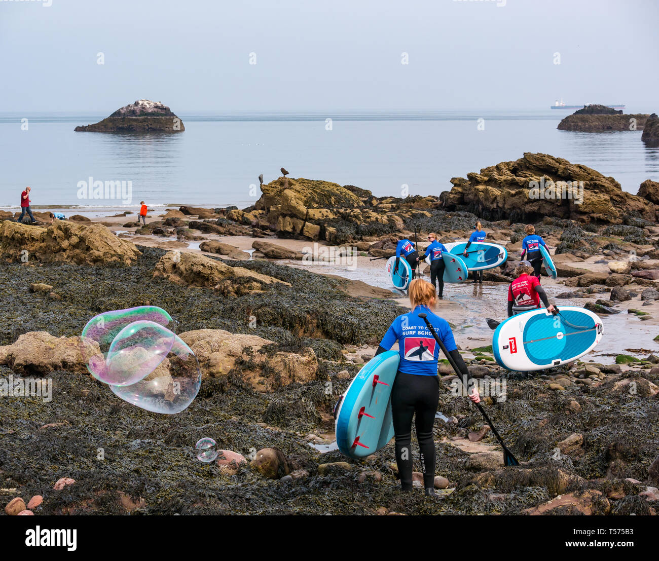 Dunbar, East Lothian, Scotland, UK. 21st Apr 2019. UK Weather:  People enjoy the very sunny hot Easter day weather at Eye Cave cove. A group of paddle boarders walk over the rocky shore to go into the sea for a paddle boarding lesson with Coast to Coast surf school Stock Photo